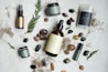 An arrangement of skincare or wellness products placed on a light surface, surrounded by natural elements such as leaves and assorted stones. The products include bottles with pump tops, jars, a spray, and a stick, each with minimalist labels in earthy tones. Dried plant material and small colorful pebbles add a rustic and organic touch.