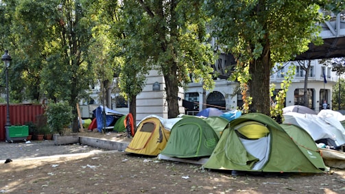 A row of colorful tents is set up in a park area surrounded by tall trees. The ground is covered with dry grass and scattered debris. In the background, urban buildings are visible, and a metal structure resembling a bridge or elevated railway can be seen.