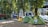 A row of colorful tents is set up in a park area surrounded by tall trees. The ground is covered with dry grass and scattered debris. In the background, urban buildings are visible, and a metal structure resembling a bridge or elevated railway can be seen.