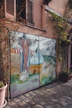 A painted mural on a garage door features a whimsical character with a long red beard and striped clothing. The scene includes mountains in the background, a body of water, and various abstract elements. The mural is framed by vines growing around it, set against a stucco building with closed wooden shutters above.