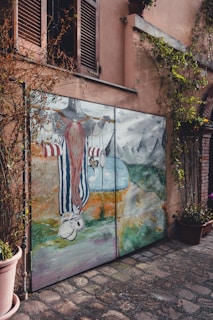 A painted mural on a garage door features a whimsical character with a long red beard and striped clothing. The scene includes mountains in the background, a body of water, and various abstract elements. The mural is framed by vines growing around it, set against a stucco building with closed wooden shutters above.