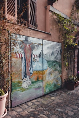 A painted mural on a garage door features a whimsical character with a long red beard and striped clothing. The scene includes mountains in the background, a body of water, and various abstract elements. The mural is framed by vines growing around it, set against a stucco building with closed wooden shutters above.