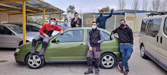 A group of seven people wearing face masks, casually posing around a green car parked in a lot. Some are leaning against the car, while one person sits on the hood. The background features additional parked cars, a yellow canopy, metal railings, and tall trees without leaves.