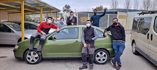 A group of seven people wearing face masks, casually posing around a green car parked in a lot. Some are leaning against the car, while one person sits on the hood. The background features additional parked cars, a yellow canopy, metal railings, and tall trees without leaves.