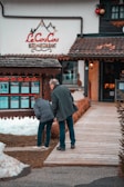 A man and a woman are looking at a display board outside a hotel and restaurant named 'Le CouCou'. They are standing on a wooden walkway surrounded by snow and a landscaped area with shrubs. The hotel's exterior features a rustic style with decorative elements.