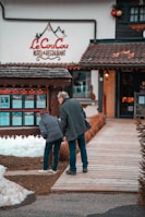 A man and a woman are looking at a display board outside a hotel and restaurant named 'Le CouCou'. They are standing on a wooden walkway surrounded by snow and a landscaped area with shrubs. The hotel's exterior features a rustic style with decorative elements.