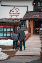 A man and a woman are looking at a display board outside a hotel and restaurant named 'Le CouCou'. They are standing on a wooden walkway surrounded by snow and a landscaped area with shrubs. The hotel's exterior features a rustic style with decorative elements.