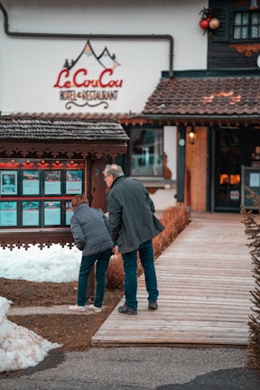 A man and a woman are looking at a display board outside a hotel and restaurant named 'Le CouCou'. They are standing on a wooden walkway surrounded by snow and a landscaped area with shrubs. The hotel's exterior features a rustic style with decorative elements.