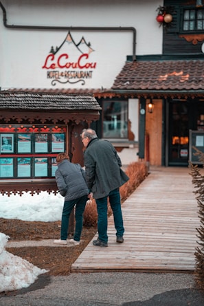 A man and a woman are looking at a display board outside a hotel and restaurant named 'Le CouCou'. They are standing on a wooden walkway surrounded by snow and a landscaped area with shrubs. The hotel's exterior features a rustic style with decorative elements.