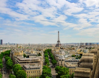 A panoramic view of Paris featuring the Eiffel Tower prominently rising in the center amidst a sprawling urban landscape. The streets are lined with lush green trees and architecturally exquisite buildings. The sky is partly cloudy, adding a picturesque backdrop to the cityscape.