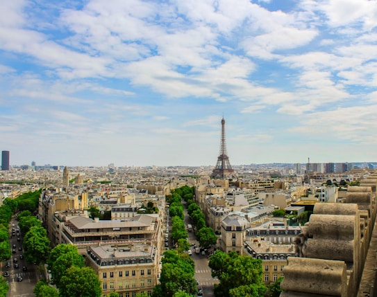 A panoramic view of Paris featuring the Eiffel Tower prominently rising in the center amidst a sprawling urban landscape. The streets are lined with lush green trees and architecturally exquisite buildings. The sky is partly cloudy, adding a picturesque backdrop to the cityscape.