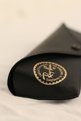 A close-up view of a black leather case with a circular gold and black logo printed on it, indicating it's a Ray-Ban sunglasses case. The focus is on the stitching and texture of the material, which is clearly visible, while the background appears blurred.