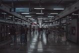 A dimly lit transit station with signs hanging from the ceiling displaying transportation information. People are walking along a platform with glass walls and doors indicating a modern setting. Fluorescent lights line the ceiling, reflecting on the polished floor.