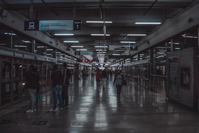 A dimly lit transit station with signs hanging from the ceiling displaying transportation information. People are walking along a platform with glass walls and doors indicating a modern setting. Fluorescent lights line the ceiling, reflecting on the polished floor.