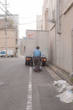 A person wearing a blue shirt and dark pants is pushing a cart towards a delivery truck in a narrow alley. The scene is urban with buildings on either side, along with overhead wires. Bags are placed on the sidewalk nearby, and the truck has logos and license plate numbers visible.