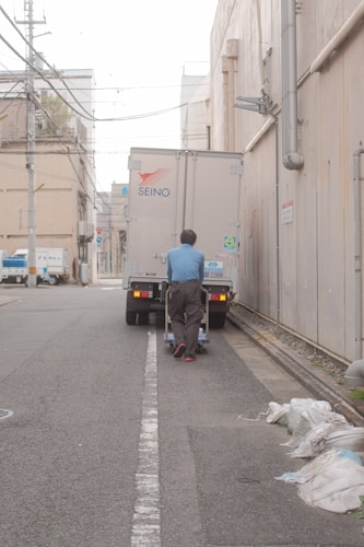 A person wearing a blue shirt and dark pants is pushing a cart towards a delivery truck in a narrow alley. The scene is urban with buildings on either side, along with overhead wires. Bags are placed on the sidewalk nearby, and the truck has logos and license plate numbers visible.