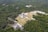 An aerial view of an ancient archaeological site surrounded by dense green forest. A large stepped pyramid structure is centrally located, with other smaller stone buildings and ruins scattered around. The area is bustling with numerous visitors exploring the site.