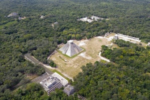An aerial view of an ancient archaeological site surrounded by dense green forest. A large stepped pyramid structure is centrally located, with other smaller stone buildings and ruins scattered around. The area is bustling with numerous visitors exploring the site.