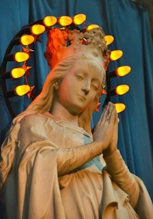 A statue of a woman is depicted in a prayerful pose with hands clasped. She wears a crown encircled by illuminated orange lights shaped like stars. The statue is set against a blue draped background, and the lighting casts a warm glow on the figure's face and robe.