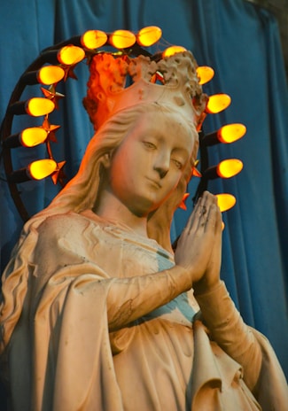 A statue of a woman is depicted in a prayerful pose with hands clasped. She wears a crown encircled by illuminated orange lights shaped like stars. The statue is set against a blue draped background, and the lighting casts a warm glow on the figure's face and robe.