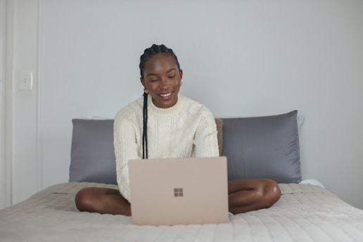 A person with braided hair, wearing a cream-colored sweater, sits cross-legged on a bed, using a laptop. The setting appears cozy with a simple, neutral color palette. The individual is smiling, creating a warm and inviting atmosphere.