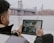 A person is sitting outdoors near a waterfront, holding a tablet in their hands. The tablet displays a video call with multiple participants. In the background, there is a large suspension bridge and a city skyline shrouded in mist.