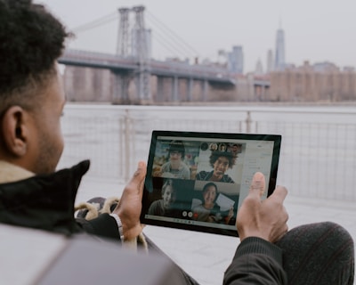 A person is sitting outdoors near a waterfront, holding a tablet in their hands. The tablet displays a video call with multiple participants. In the background, there is a large suspension bridge and a city skyline shrouded in mist.