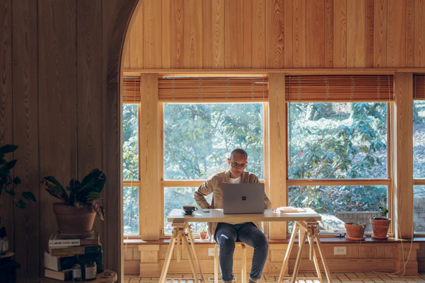 Stephanie Shaw writing at a rustic wooden desk by a window overlooking a peaceful garden filled with ferns and leafy plants.