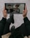 A person is holding a tablet while sitting, participating in a video conference call with four people displayed on the screen. The screen also shows a chat section with messages and emojis.