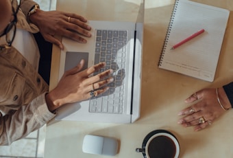 Two individuals are working at a desk. One person's hands are typing on a laptop, and the other person's hands are resting on the table, with both wearing rings. There is an open notebook with a red pen on top, a white mouse, and a cup of coffee nearby.