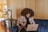 A person with curly hair is sitting on a blue couch, engaged in a video call on a laptop. The setting appears to be a cozy, wood-paneled room with a television, potted plants, and warm lighting.