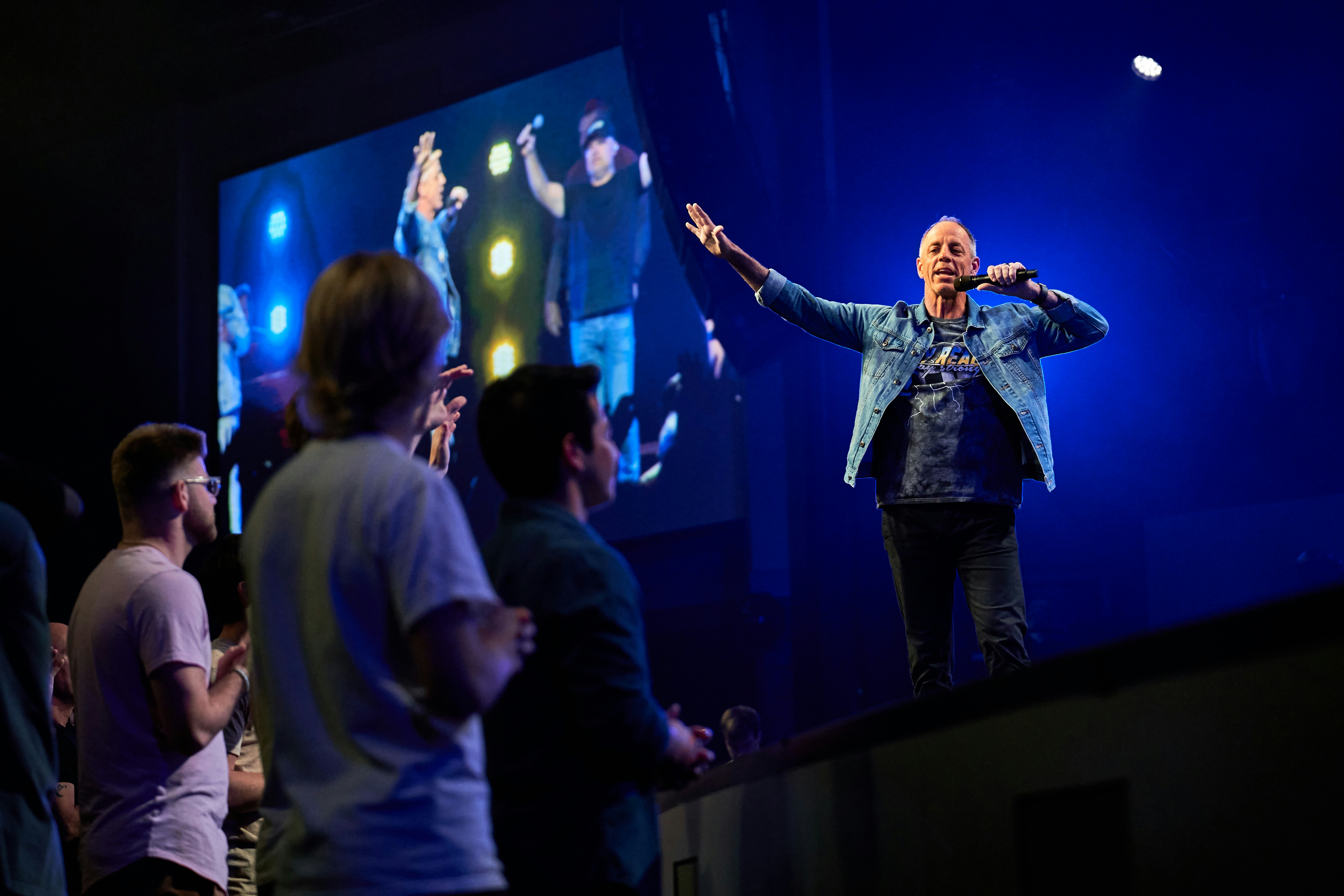 man on a stage singing with a video screen behind him and a crowd looking on