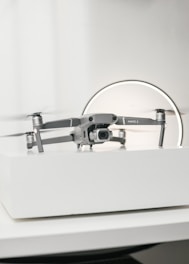 A compact, silver drone sits on a white surface, showcasing a modern and sleek design with its camera facing forward. The background features a circular light, giving the scene a subtle futuristic vibe.