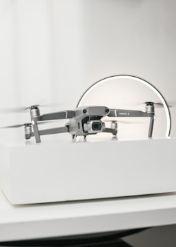 A compact, silver drone sits on a white surface, showcasing a modern and sleek design with its camera facing forward. The background features a circular light, giving the scene a subtle futuristic vibe.