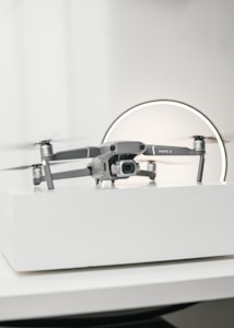A compact, silver drone sits on a white surface, showcasing a modern and sleek design with its camera facing forward. The background features a circular light, giving the scene a subtle futuristic vibe.
