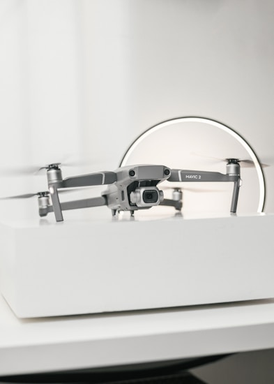 A compact, silver drone sits on a white surface, showcasing a modern and sleek design with its camera facing forward. The background features a circular light, giving the scene a subtle futuristic vibe.