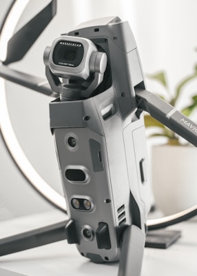 A detailed close-up of a high-tech drone, featuring a prominent Hasselblad camera with a wide-angle lens. The design is sleek and modern with a metallic finish, and the drone appears to be set on a table with a blurred background.
