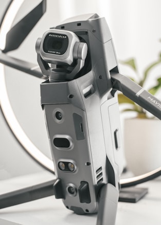 A detailed close-up of a high-tech drone, featuring a prominent Hasselblad camera with a wide-angle lens. The design is sleek and modern with a metallic finish, and the drone appears to be set on a table with a blurred background.