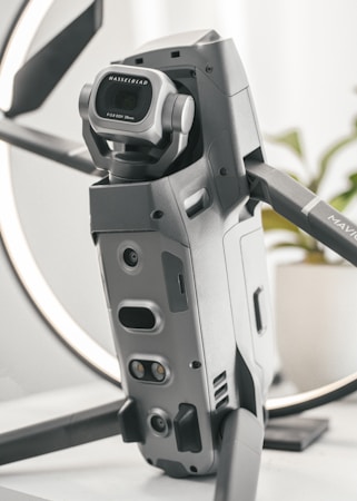 A detailed close-up of a high-tech drone, featuring a prominent Hasselblad camera with a wide-angle lens. The design is sleek and modern with a metallic finish, and the drone appears to be set on a table with a blurred background.