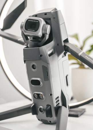 A detailed close-up of a high-tech drone, featuring a prominent Hasselblad camera with a wide-angle lens. The design is sleek and modern with a metallic finish, and the drone appears to be set on a table with a blurred background.