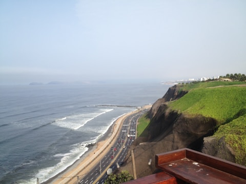 A coastal landscape featuring a winding road along a cliffside that overlooks the ocean. The road is busy with traffic and runs parallel to sandy beaches with waves crashing on the shore. On the right, lush green hills rise steeply, contrasting with the vast expanse of the water. Buildings can be seen in the distance, blending into the horizon.