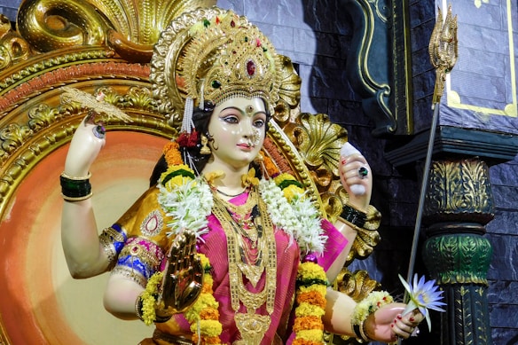 A detailed and ornate statue dressed in vibrant clothing and adorned with elaborate jewelry. The figure holds various items in her hands, including a lotus flower and a discus. Garlands of flowers drape over her shoulders, and she is set against a backdrop of rich textures and colors.