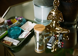 On a reflective glass surface, there are items commonly associated with relaxation and leisure. A golden, stylized statue shaped like a person in a meditative pose stands beside a collection of smoking accessories, including a pipe, rolling papers, lighters, and containers. There are also small jars with wooden lids containing dried plant material, and a candle set in a metal container.