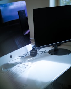 A modern workspace is set up with a sleek design, featuring a large Apple iMac with a visible logo and a black monitor on a white desk. Cables are neatly organized behind the screen, and a lens is placed between the monitors. Natural light filters in through a window, creating a soft illumination across the surfaces.