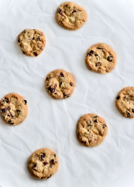 A collection of eight freshly baked cookies with chocolate chips, arranged in a random pattern on a crinkled white parchment paper. The cookies are golden brown with a soft texture and the chocolate chips are visibly embedded.