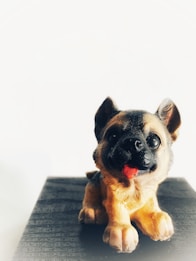 A small figurine of a German Shepherd puppy is depicted, positioned on a matte black surface against a white background. The puppy's mouth is open with a pink tongue visible, and it features detailed fur with shades of black and tan.
