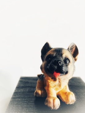 A small figurine of a German Shepherd puppy is depicted, positioned on a matte black surface against a white background. The puppy's mouth is open with a pink tongue visible, and it features detailed fur with shades of black and tan.