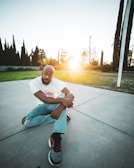 A person sits on a concrete surface in an outdoor setting. They are wearing a white t-shirt with the word 'Thirdlife' and light blue jeans with rips at the knees. The person is gazing towards the camera with a thoughtful expression. The sun is setting in the background, casting a warm glow across the scene. Tall trees and a grassy area surround the paved space.