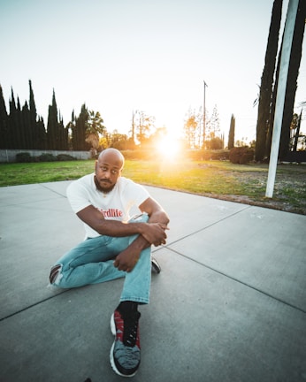 A person sits on a concrete surface in an outdoor setting. They are wearing a white t-shirt with the word 'Thirdlife' and light blue jeans with rips at the knees. The person is gazing towards the camera with a thoughtful expression. The sun is setting in the background, casting a warm glow across the scene. Tall trees and a grassy area surround the paved space.