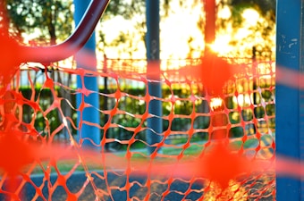 Bright orange netting is prominently featured in the foreground, with sunlight streaming through it, casting intricate patterns. Metal poles frame the net, and blurred greenery is visible in the background.